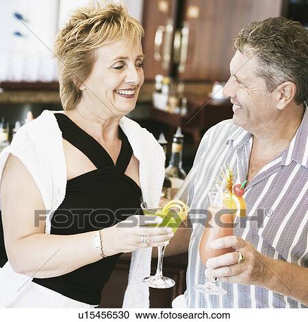 Stock Image - Senior man and a mature woman holding glasses of cocktail and standing at a bar counter. Fotosearch