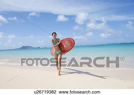 Stock Image - A young woman holding surfboard at beach. Fotosearch