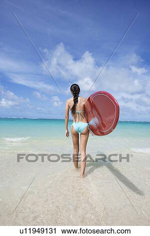 Stock Image - Rear view of a woman holding surfboard at beach. Fotosearch
