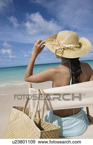Stock Photo - Rear view of a woman sitting on chair at beach. Fotosearch