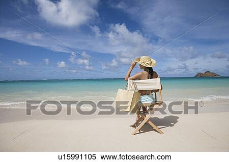 Stock Photography - Rear view of a woman sitting on chair at beach. Fotosearch