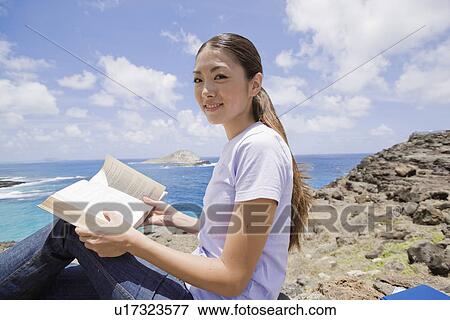 Stock Photo - View of a young woman sitting on rock with book. Fotosearch