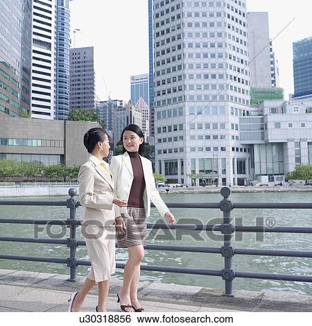 Stock Photograph - Side profile of two businesswomen walking on a walkway. Fotosearch