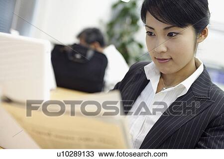 Stock Image - A businesswoman interestingly reading the newspaper with a laptop placed beside her. Fotosearch