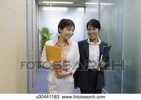 Stock Image - Two professional women holding files walks along the passage of the office. Fotosearch