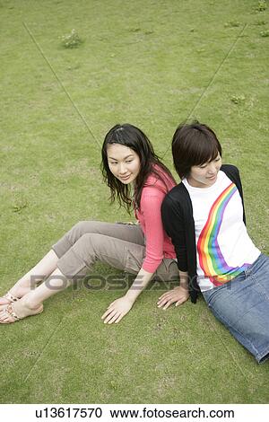 Stock Image - Two young women rest on each others back as they sit on the grassland. Fotosearch