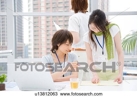 Stock Image - Two young women looking at notebook and young man standing behind. Fotosearch