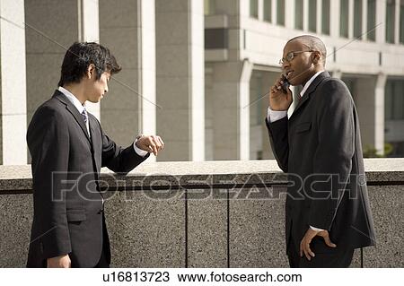 Stock Image - Two businessmen, one using mobile phone. Fotosearch