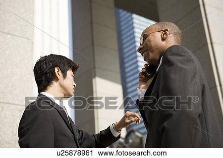 Stock Image - Two businessmen, one using mobile phone. Fotosearch