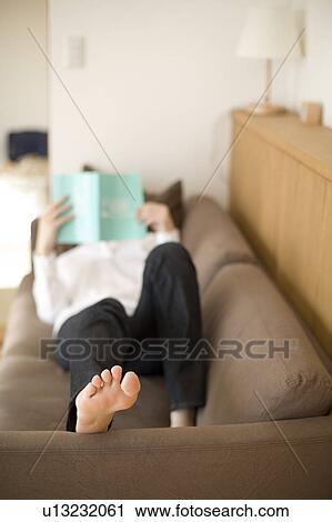 Stock Image - Young woman reading book lying on sofa. Fotosearch