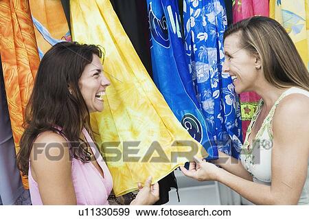 Stock Photo - Close-up of two mid adult women looking clothes at a market stall and smiling. Fotosearch