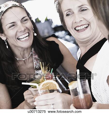 Stock Photography - Mid adult woman and a mature woman holding glasses of cocktail and smiling. Fotosearch