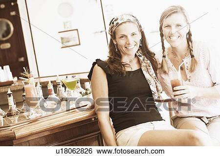 Stock Photograph - Portrait of two mid adult women sitting at a bar counter and smiling. Fotosearch