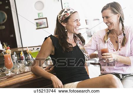 Picture - Two mid adult women sitting at a bar counter and smiling. Fotosearch