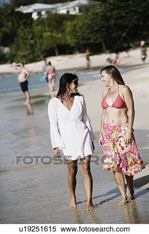 Stock Photography - Two mid adult women walking on the beach with tourists in the background. Fotosearch