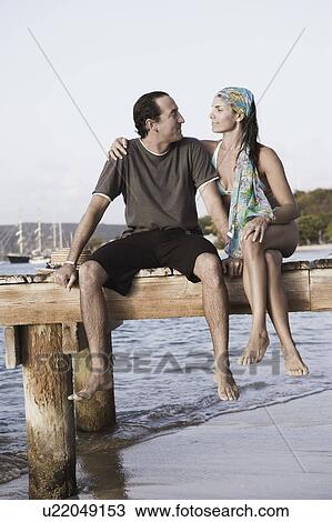 Mid adult woman with her arm around a mid adult man sitting on a jetty View Large Photo Image Stock Image - Mid adult woman with her arm around a mid adult man sitting on a jetty. Fotosearch
