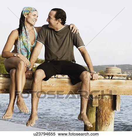 Stock Photography - Mid adult woman with her arm around a mid adult man sitting on a jetty. Fotosearch
