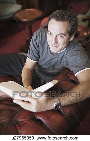 Stock Image - High angle view of a mid adult man sitting on a couch and holding a book. Fotosearch