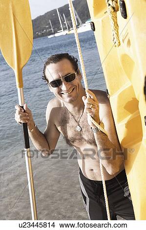 Picture - Mid adult man standing with an oar and a surfboard on the beach. Fotosearch