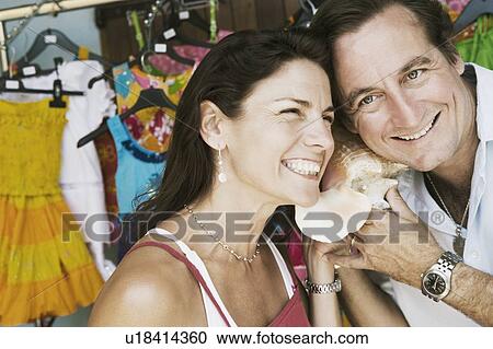Stock Image - Close-up of a mid adult couple listening to a conch shell and smiling. Fotosearch