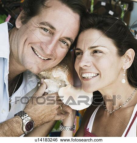 Stock Photograph - Close-up of a mid adult couple listening to a conch shell and smiling. Fotosearch