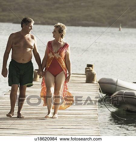 Picture - Senior man and a mature woman holding each other's hands and walking on a jetty. Fotosearch
