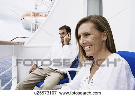 Stock Photo - Mid adult man talking on a mobile phone with a mid adult woman sitting on a chair and smiling. Fotosearch