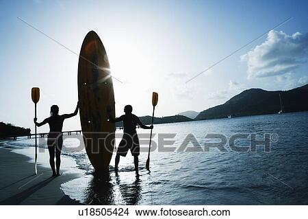 Picture - Silhouette of a man and a woman holding oars and a surfboard on the beach. Fotosearch