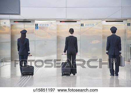Stock Photo - Rear view of a male and a female pilot walking with a female cabin crew in front of elevators. Fotosearch