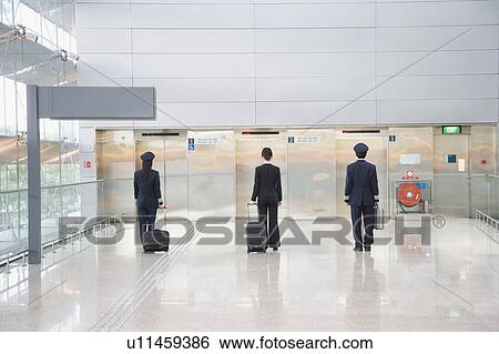 Stock Photograph - Rear view of a male and a female pilot walking with a female cabin crew in front of elevators. Fotosearch