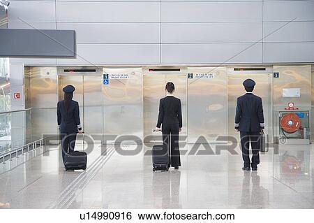 Stock Photograph - Rear view of a male and a female pilot walking with a female cabin crew in front of elevators. Fotosearch