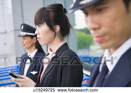 Stock Photography - Side profile of a female cabin crew holding a mobile phone and standing with two pilots at an airport. Fotosearch