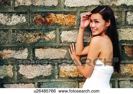Stock Photograph - Young woman leaning on wall, smiling, side view. Fotosearch