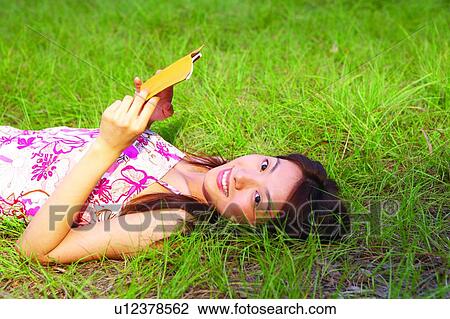 Stock Image - Young woman lying on grass, holding a notepad. Fotosearch