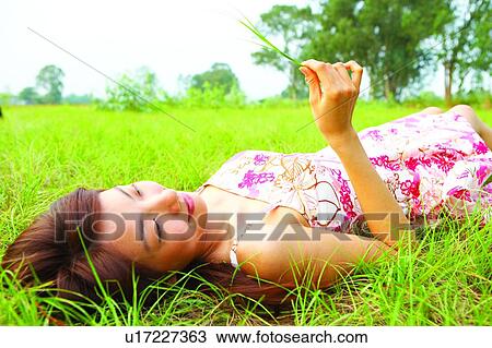 Stock Image - Young woman lying on grass, holding a long grass. Fotosearch