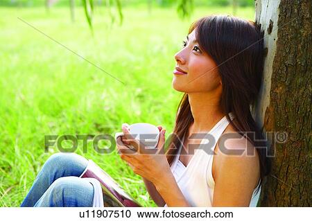 Young woman sitting on grass and leaning to tree, holding a cup, close-up View Large Photo Image Stock Image - Young woman sitting on grass and leaning to tree, holding a cup, close-up. Fotosearch