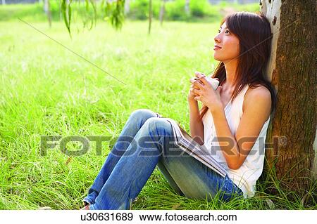 Stock Photo - Young woman sitting on grass and leaning to tree, holding a cup. Fotosearch