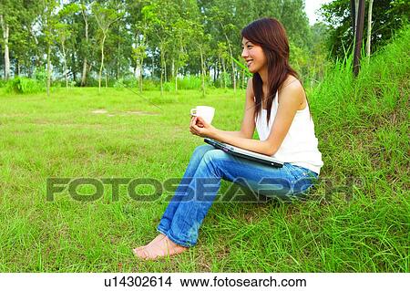 Picture - Young woman sitting on grass, holding a cup, full length. Fotosearch