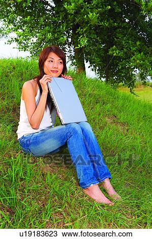 Stock Image - Young woman sitting on grass, holding a notebook, full length. Fotosearch