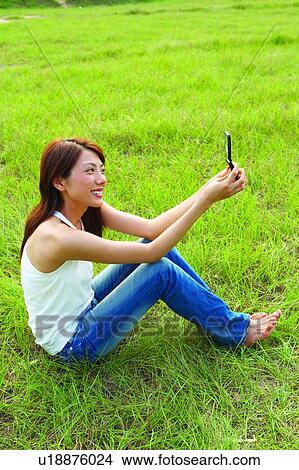 Young woman sitting on grass, holding mobile phone, cheerful, bending legs View Large Photo Image Picture - Young woman sitting on grass, holding mobile phone, cheerful, bending legs. Fotosearch