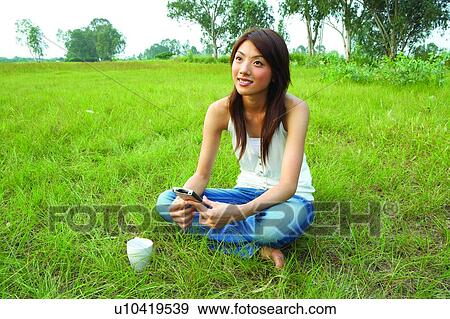 Stock Photo - Young woman sitting on grass, holding pda. Fotosearch