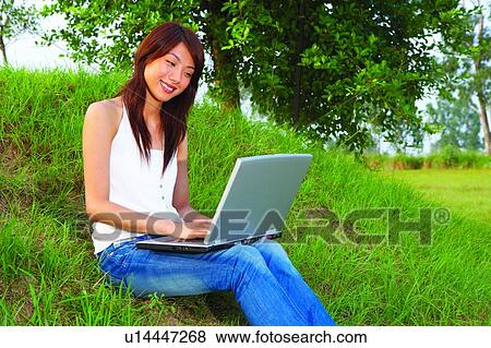 Stock Photo - Young woman sitting on grass, using notebook computer. Fotosearch