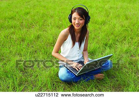 Young woman sitting on grass, wearing headphone, using notebook computer View Large Photo Image Stock Image - Young woman sitting on grass, wearing headphone, using notebook computer. Fotosearch