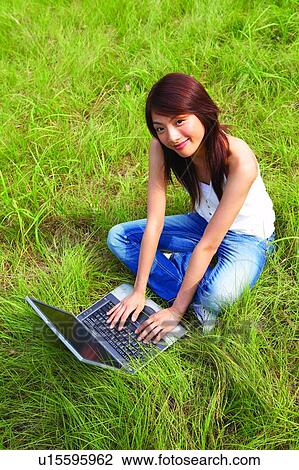 Stock Image - Young woman sitting on green grass, using notebook computer. Fotosearch