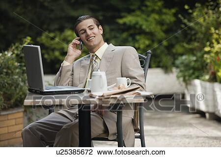 Stock Image - Businessman in garden cafe with cell phone and laptop. Fotosearch