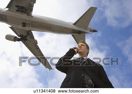Stock Photo - A Businessman Talking on a Cell Phone with an Airplane in the Background, Low Angle View, Waist Up. Fotosearch