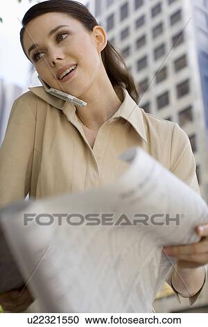 Stock Image - A Young Adult Businesswoman Holding a Newspaper and Talking on a Cellular Phone, Low Angle View, Front View, Waist Up, Differential Focus. Fotosearch