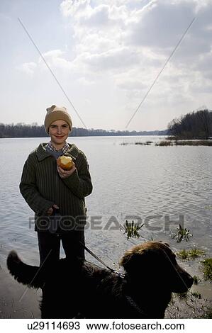 Stock Image - Boy (12-14) walking dog by river, holding apple, smiling, portrait. Fotosearch