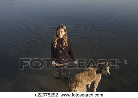 Stock Image - Girl (10-12) standing in river holding dog on lead, smiling, portrait. Fotosearch