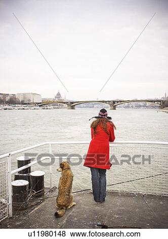 Stock Photo - Hungary, River Danube, young woman on jetty with dog, rear view. Fotosearch